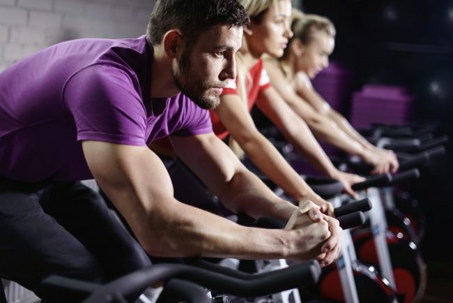Close up hands of man biking in spinning class. Group of smiling friends at gym exercising on stationary bike. Happy cheerful athletes training on exercise bike.